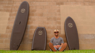 Man sitting along a wall wearing glasses next to 3 surfboards