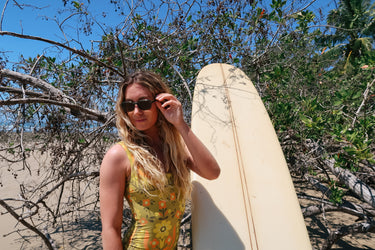 Woman holding a surfboard in a natural setting with trees and sand.